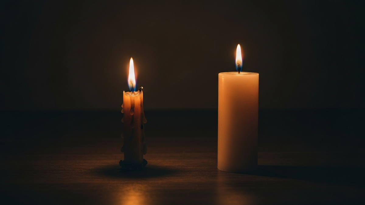 A worn burned-down candle glowing beside a fresh unlit candle on a dark wooden surface