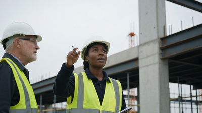 A civil engineer in a hard hat on a construction site, looking up at unfinished steelwork with a colleague.