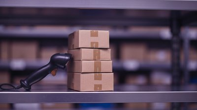 Handheld inventory scanner beside a stack of cardboard boxes on a retail stockroom shelf, purple-toned ambient light