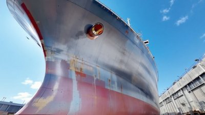The grey and red painted steel hull of a large ship under construction in a sunny dry dock