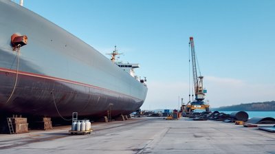 The curved grey steel hull of a large ship resting on keel blocks beside a concrete repair quay and a tall crane