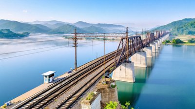 A steel railway truss bridge with double tracks crossing a wide river between green hills